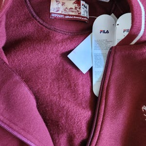 May include: A burgundy FILA zip-up sweatshirt with a white stripe detail on the collar. The sweatshirt has a Ferrari official licensed product label and a FILA tag attached. The zipper is visible, and the fabric appears soft.