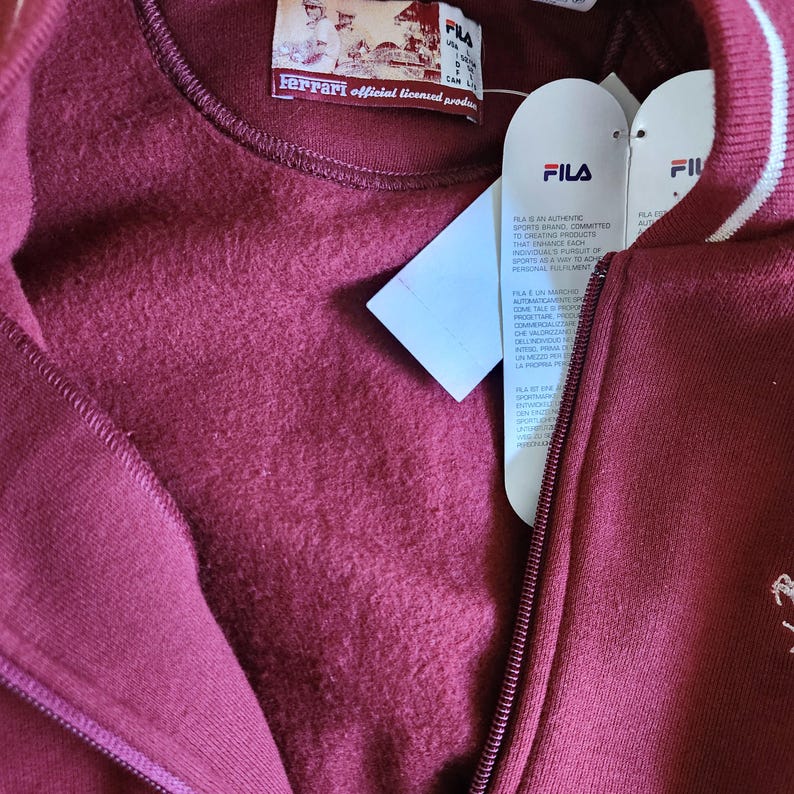 May include: A burgundy FILA zip-up sweatshirt with a white stripe detail on the collar. The sweatshirt has a Ferrari official licensed product label and a FILA tag attached. The zipper is visible, and the fabric appears soft.
