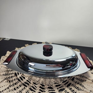 May include: A chrome serving dish with a domed lid and two dark red handles. The lid has a matching red knob. The oval-shaped dish sits on a cream-coloured, open-weave doily.