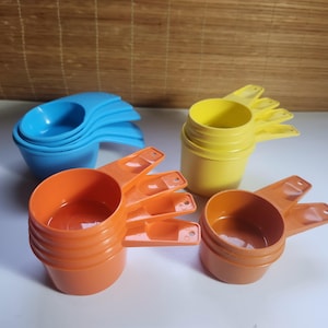 Könnte beinhalten: Eine Sammlung farbiger Messbecher in Blau, Gelb und Orange. Die Becher sind in Sets gestapelt, mit Griffen. Das Bild zeigt eine Vielzahl von Größen, geeignet für die Küche. Maße in metrischen Einheiten.
