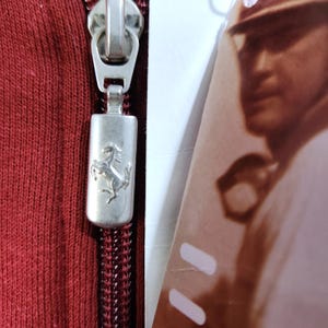 May include: Close-up of a red zipper with a silver pull featuring the Ferrari logo, a prancing horse. The zipper is on a red fabric, and a portion of a tag with a blurred image is visible.