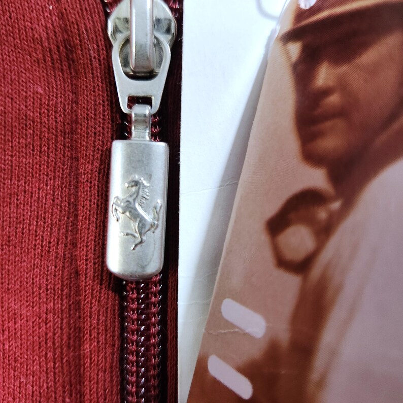 May include: Close-up of a red zipper with a silver pull featuring the Ferrari logo, a prancing horse. The zipper is on a red fabric, and a portion of a tag with a blurred image is visible.