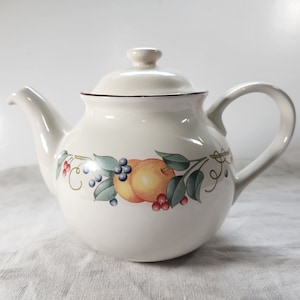 Op de afbeelding: Crèmekleurige keramische theepot met deksel en handvat. De theepot is versierd met een fruit- en bladontwerp met perziken, bosbessen en rode bessen. De rand is afgewerkt met een donkerbruine lijn.