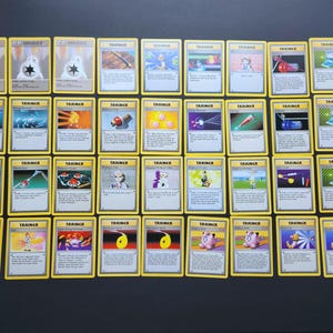 Puede incluir: Una colección de cuarenta cartas coleccionables vintage dispuestas en una cuadrícula, que presentan varias cartas de Energía y Entrenador con bordes amarillos, cuadros de texto blancos e ilustraciones coloridas de objetos, personajes y símbolos.