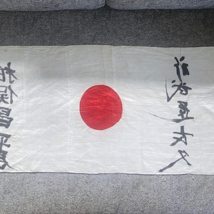 May include: A white rectangular flag with a large red circle in the centre, resembling the Japanese flag. Black Japanese characters are written on the flag. The flag is lying on a grey surface.