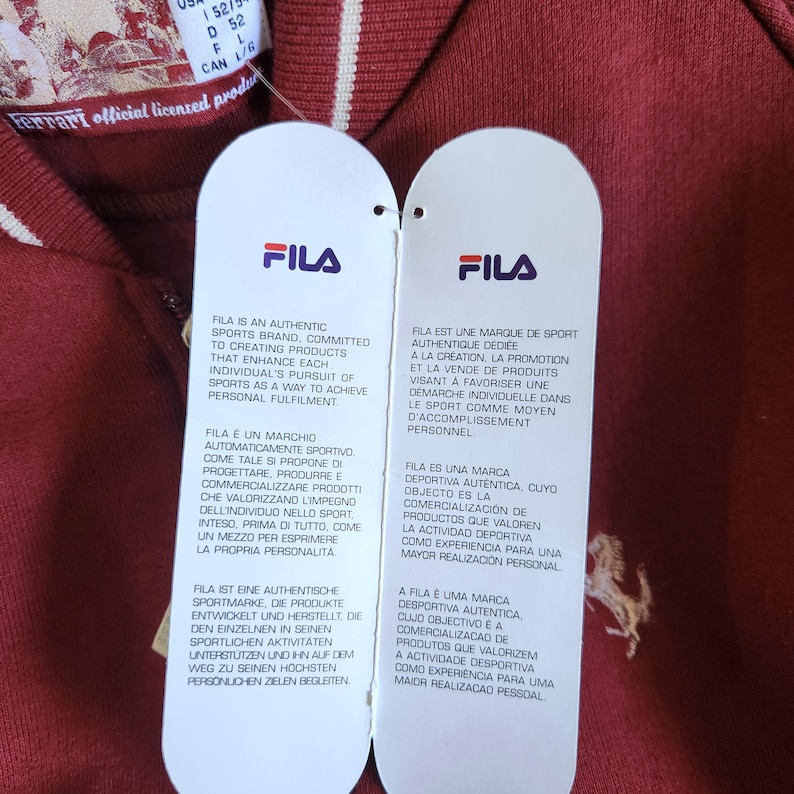 May include: Close-up of a burgundy Fila polo shirt with white stripes on the collar and sleeves. Two white tags with the Fila logo and brand information are attached. The tags have text in multiple languages.