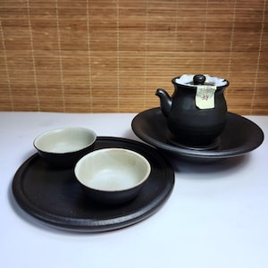 Puede incluir: Un juego de té de cerámica negra con una tetera, dos cuencos pequeños y dos platos. La tetera tiene tapa y boquilla. Los cuencos tienen un interior de color claro. El juego está sobre una superficie blanca con un fondo de persiana de bambú.