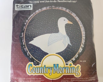 May include: A craft kit featuring a white goose with a blue and white checked scarf. The kit includes fabric, trims, and patterns. The packaging has the text "Country Morning" and "An easy and fun-to-do Needlecraft kit".