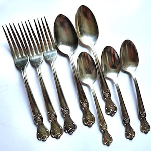 May include: A collection of vintage silverware, including forks and spoons, arranged on a white surface. The silverware has ornate floral detailing on the handles and a shiny, reflective finish. The set includes both large and small spoons.