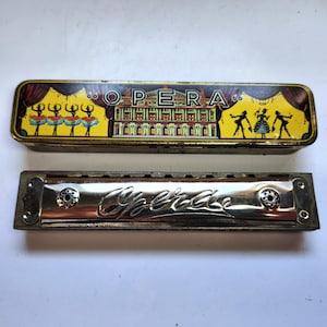May include: A vintage harmonica and its rectangular metal storage case. The case features a yellow background with a colorful illustration of an opera house, dancers, and the text "OPERA". The silver-toned harmonica has the word "Opera" embossed.