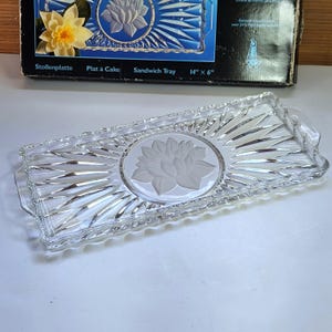 May include: A rectangular clear glass tray featuring a central frosted lotus flower motif surrounded by a sunburst pattern of radiating ridges. The tray has integrated handles on both ends and measures 14 inches by 6 inches as stated on the box.