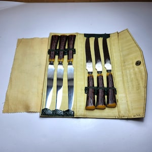以下が含まれることがあります： 質感のある茶色のハンドルと銀色の金属製ブレードを備えた6本のステーキナイフセット。淡い黄色の内装とダークグリーンの質感のあるトリムが付いた折りたたみ式ケースに収納されています。ナイフは3本ずつ2列に並べられています。