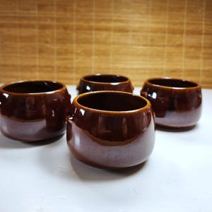 May include: Four brown ceramic bowls with a glossy finish. The bowls have a rounded shape with a slightly flared rim and small handles. The interior of the bowls is a darker brown color.