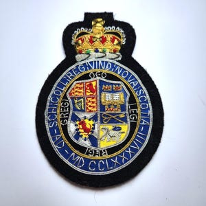 May include: A circular embroidered patch featuring a gold crown at the top and a central shield divided into four quadrants with heraldic symbols. The blue border contains the text SCHNCOLLI REGIVIND NOVA SCOTIA AD MDCCLXXXVIII.
