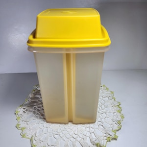 以下が含まれることがあります： 黄色いプラスチック製の容器と蓋。容器は透明で、表面にテクスチャがあります。容器は白いドイリーの上に置かれています。
