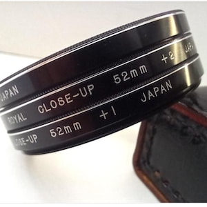 Puede incluir: Filtros de lente Royal Close-Up negros, marcados con las potencias "+1" y "+2", y tamaño "52mm". Los filtros están etiquetados como "Japan" y están apilados. Un estuche de cuero es parcialmente visible en el fondo.