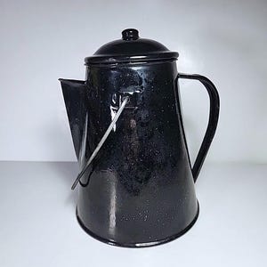 Pode incluir: Cafeteira esmaltada preta com tampa, bico e alça. A panela tem um padrão salpicado e uma alça de metal. A cafeteira é de estilo vintage e provavelmente é usada para acampar ou atividades ao ar livre.