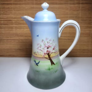 May include: A tall, ceramic coffee pot with a light blue lid and handle. The body of the pot is painted with a scene of a flowering tree, birds, and a grassy field. The handle has a gold trim.