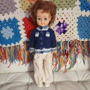 Puede incluir: Muñeca vintage con cabello castaño rojizo y ojos azules, vestida con un suéter de punto azul marino con detalles blancos y botones a juego. La muñeca también lleva pantalones color crema y zapatos blancos. El fondo presenta una colorida manta con flecos.