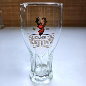 May include: A clear glass beer glass with a flared design. The glass displays the Alexander Keith's logo in gold and red, with the words "Fine Beers" beneath. The glass has a solid base and a smooth, transparent surface.