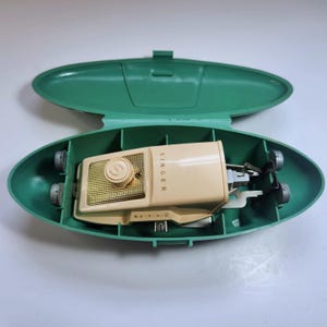 Puede incluir: Un accesorio de máquina de coser Singer de color beige alojado en un estuche de almacenamiento de plástico verde de forma ovalada. El accesorio cuenta con una placa texturizada dorada con un dial central marcado con una S, y la marca Singer está impresa en el lateral.