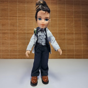 Könnte beinhalten: Eine Bratz-Puppe in formeller Kleidung. Die Puppe hat dunkles Haar, das zu einem Faux Hawk gestylt ist, und blaue Augen. Sie trägt eine hellblaue Jacke über einem gerüschten grauen Hemd, dunkelblaue Hose und braune Schuhe. Eine kleine weiße Blume ziert die Jacke.