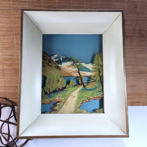May include: A rectangular shadow box lamp featuring a layered paper cutout scene of a mountain landscape with a lake, trees, and a path. The frame has a dark brown exterior and a white interior, with an attached brown electrical power cord.