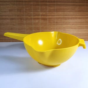 Puede incluir: Un cuenco mezclador de plástico amarillo brillante con asa y boquilla. El cuenco tiene forma redondeada con base plana y un asa pequeña en el lateral. El asa mide aproximadamente 15 cm de largo.