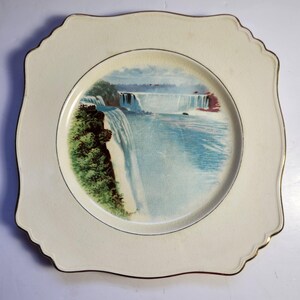 Könnte beinhalten: Cremefarbene, quadratische Zierplatte mit Goldrand. In der Mitte befindet sich ein Landschaftsbild eines Wasserfalls mit blauem Wasser, grünem Laub und einem hellblauen Himmel. Der Teller hat einen gewellten Rand.