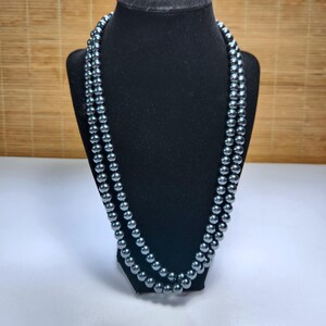May include: A long, double-strand necklace featuring uniform, round, dark metallic gray beads with a high-gloss, reflective finish. The beads are strung together in a continuous loop, creating a layered appearance around the neck display form.