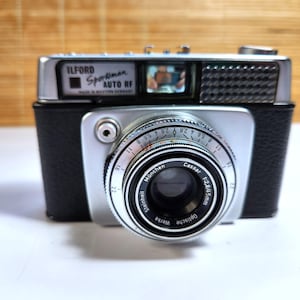 May include: An Ilford Sportsman Auto RF camera with a black textured body and silver metal top plate. The lens is a Steinheil München Cassar 1:2.8/45mm. The front features a viewfinder, a textured silver panel, and manual aperture settings.