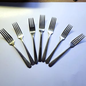 Può includere: Una collezione di sette forchette d'argento disposte a ventaglio. Le forchette hanno rebbi e lunghi manici, alcuni con motivi decorativi. Le forchette sono poste su uno sfondo bianco.