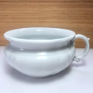 Op de afbeelding: Een witte keramische kom met een handvat. De kom heeft een brede, afgeronde vorm met een decoratief bloemmotief en een gebogen handvat. De kom staat op een wit oppervlak.