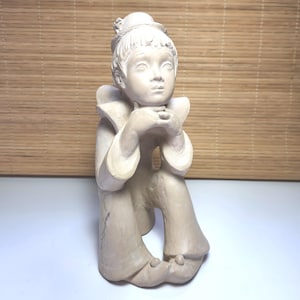 Op de afbeelding: Een beige keramieken beeldje van een zittende figuur met een hoed en een gerimpelde kraag, die haar kin op haar handen laat rusten. De figuur draagt een broek met uitlopende pijpen en kleine, afgeronde schoenen. De sculptuur heeft een matte afwerking.