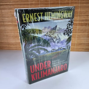 以下が含まれることがあります： アーネスト・ヘミングウェイ著「アンダー・キリマンジャロ」というタイトルのハードカバー本。表紙には山、ヤシの木、暗い背景に赤い文字でタイトルが描かれた風景が描かれています。本はビニールで密封されています。