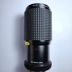 May include: A black camera lens featuring a textured, diamond-patterned grip ring. The top edge displays the text FOCAL and 55. A yellow sticker reads PASSED, and the base is marked with the white text LENS MADE IN JAPAN in a cylindrical design.