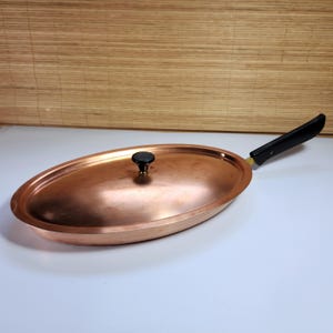 Puede incluir: Sartén ovalada de cobre con mango y tapa negros. La tapa tiene un pomo negro. La sartén es de cobre y tiene un acabado brillante. El mango es negro con un acento amarillo.