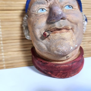 May include: A wall-mounted ceramic sculpture of a person's head wearing a blue cap and a red scarf. The figure has light blue eyes, a prominent nose, and a cigar with a glowing red tip in the mouth. The skin tone is a weathered, textured tan.