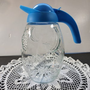 Puede incluir: Jarra de vidrio transparente con tapa y asa de plástico azul. La jarra tiene diseños de frutas en relieve, incluyendo uvas, plátanos y otras frutas. El asa y la tapa son de color azul brillante. La jarra se utiliza probablemente para servir bebidas.