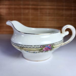 Puede incluir: Salsera de porcelana color crema con una banda floral decorativa con flores rosas y azules. El borde y el asa están adornados con ribetes dorados. La salsera tiene un pico curvo y un asa enrollada.