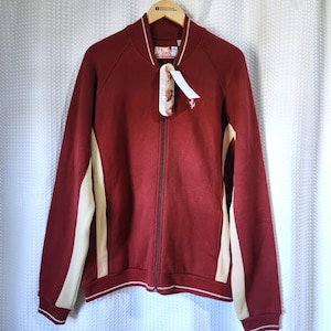 May include: A burgundy zip-up jacket with cream-colored side panels and white trim. The jacket has a ribbed collar and cuffs. A tag hangs from the zipper. The jacket is hanging on a wooden hanger.