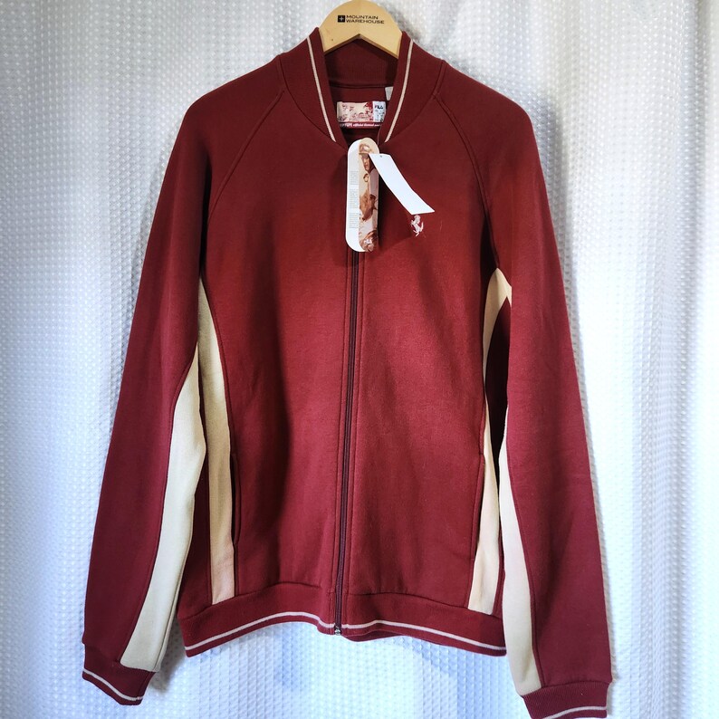 May include: A burgundy zip-up jacket with cream-colored side panels and white trim. The jacket has a ribbed collar and cuffs. A tag hangs from the zipper. The jacket is hanging on a wooden hanger.