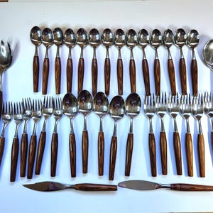 Può includere: Un set di posate in acciaio inossidabile con manici in legno. Il set comprende cucchiai, forchette e coltelli, disposti su una superficie bianca. I manici in legno sono di colore marrone scuro.