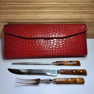 Può includere: Una custodia per coltelli in pelle rossa strutturata con un set da intaglio abbinato. Il set include un coltello da intaglio, una forchetta da intaglio e un affilatore. I manici hanno un motivo marrone e giallo.