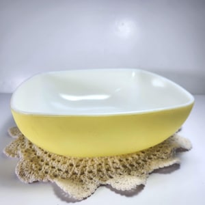 May include: A vintage, square-shaped bowl with rounded corners. The exterior is a soft yellow, while the interior is white. It rests on a beige, crocheted doily. A classic piece for serving or display.