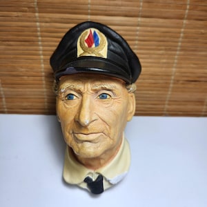 May include: A ceramic bust of an older individual wearing a black captain's hat with a gold emblem featuring red and blue flags. The figure has light skin, blue eyes, and a neutral expression, dressed in a cream-colored collared shirt with a black tie.