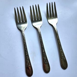 May include: Three silver forks with ornate floral detailing on the handles. The tines are evenly spaced. The forks are arranged on a white surface, showcasing their elegant design. A classic set for dining.