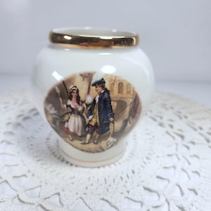 May include: A small, white ceramic jar with a gold rim. The jar features a painted scene of two figures in period clothing, with a bridge and carriage in the background. The jar is sitting on a white, lacy surface.