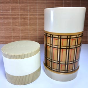 Puede incluir: Juego de termo y recipiente Aladdin vintage. El termo tiene un patrón de cuadros marrón, rojo y negro en la mitad inferior, con una parte superior blanca. El recipiente es blanco con una base y tapa beige, con el logotipo de Aladdin.