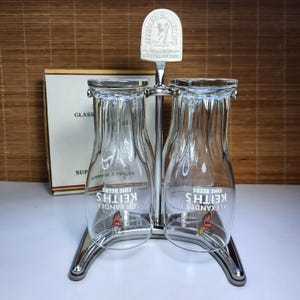 May include: A pair of clear glass beer glasses, each with "Alexander Keith's Fine Beers" printed on them, held in a chrome metal stand. The stand has a decorative top with the Alexander Keith's logo. A box is in the background.